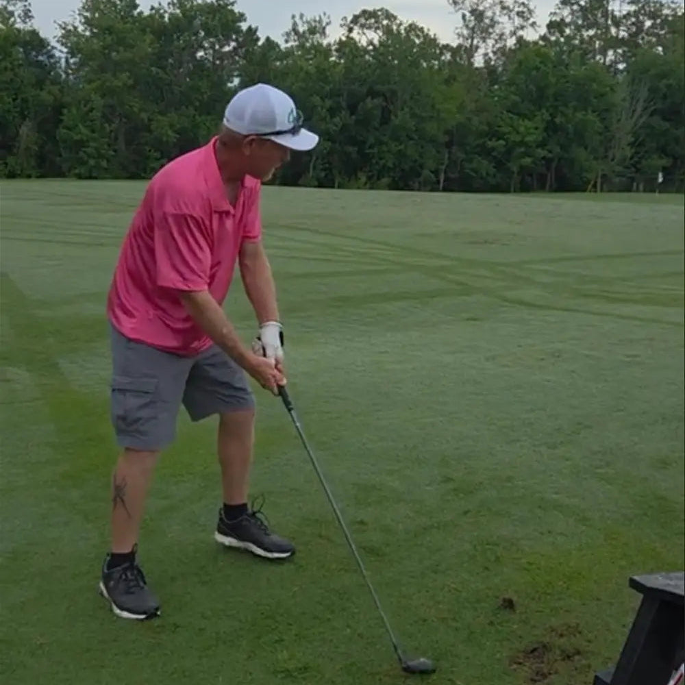 Man in pink shirt and Ferret Script Hat playing golf on a green course with trees in the background
