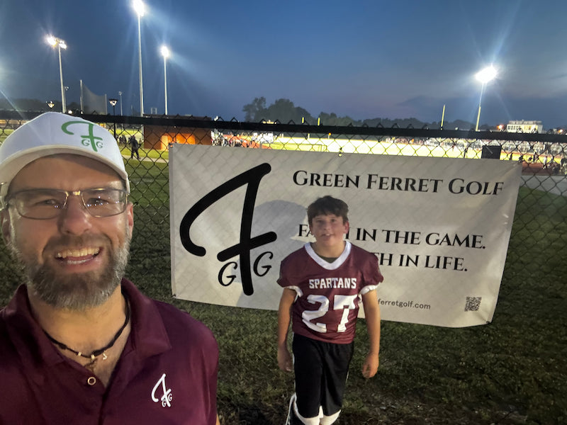Rob in a maroon Pro Shop polo with Zach standing in front of a 'Green Ferret Golf' banner at the Sun Valley Spartans High School football stadium in Indian Trail, NC
